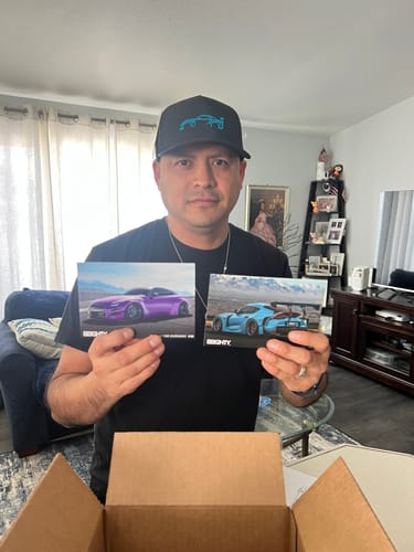Customer wearing the black 80Eighty® Limited Edition DCG87 Hat while holding up two photos of cars.