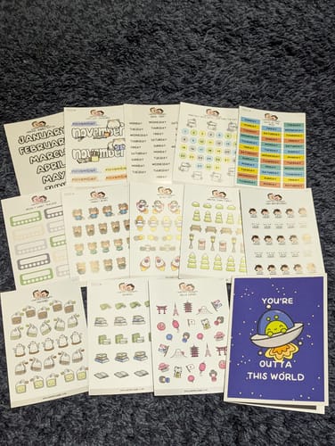 Reviewer's assortment of planner and character sticker sheets from an Oopsie Mystery Bag laid out on a dark surface.
