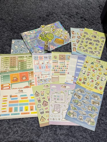 Reviewer's collection of colorful sticker sheets and a holographic card from an Oopsie Mystery Bag on a dark surface.