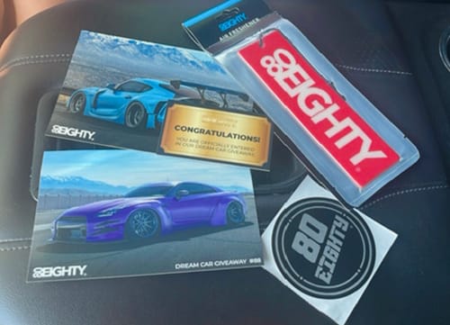 Customer's packaged red 80Eighty Air Freshener displayed on a car seat with promotional flyers and a sticker.