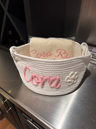 Customer's personalized white knitted basket with 'Cora' in pink yarn and a flower icon, holding a matching blanket.