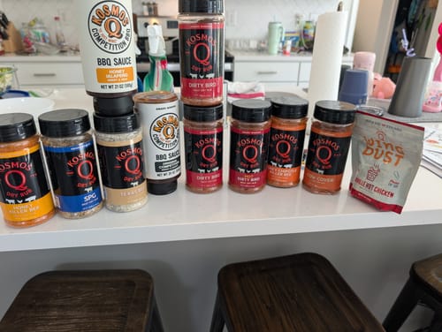 Reviewer's collection of Kosmos Q rubs and sauces from the Pit Master Gift Basket, displayed on a kitchen counter.