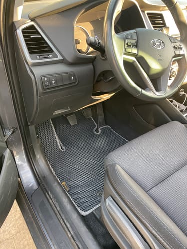 Recenzent pokazuje dopasowany szary dywanik samochodowy po stronie kierowcy.