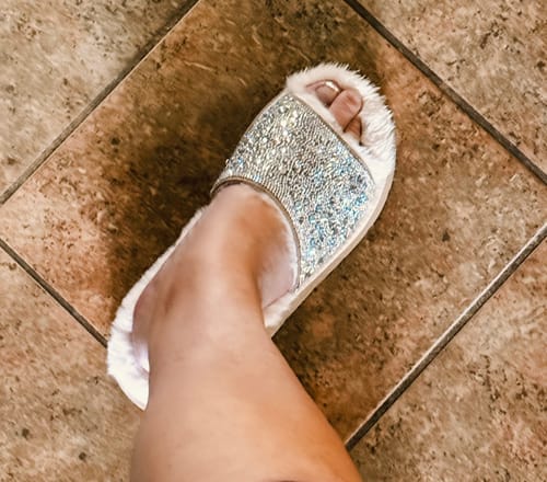 Reviewer's foot in a fuzzy Corky's shoe with a sparkling rhinestone strap, seen from above on a tile floor.