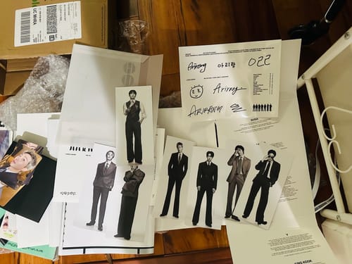 Reviewer's BTS - ARIRANG 5TH ALBUM unboxed, showing photocards and other paper inclusions on a wooden floor.