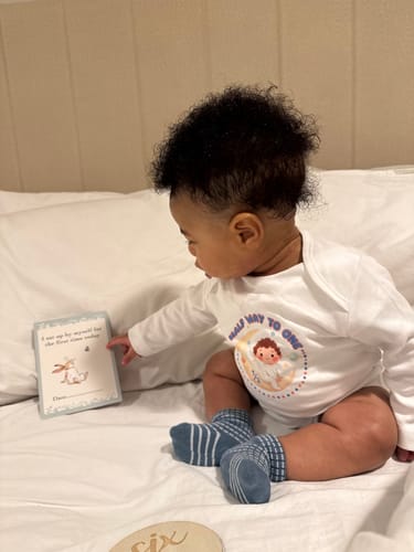 A reviewer's baby sitting on a bed wearing the white long sleeve bodysuit with a custom 'Half way to one' graphic.