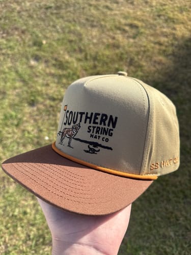Reviewer holds the tan and brown Hat of the Month Crew cap outdoors, showing the front logo and rope detail.