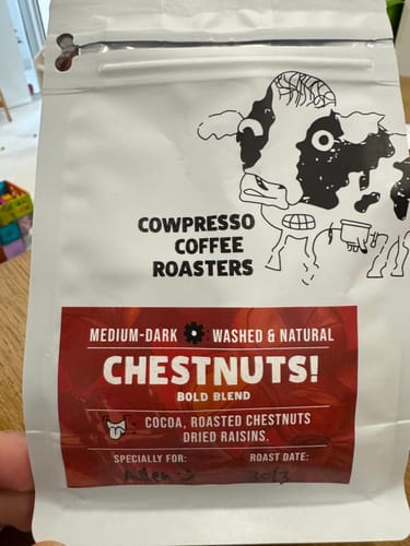Reviewer holding a white bag of Cowpresso "Chestnuts! Bold Blend" coffee from their Weekly Surprise Plan.