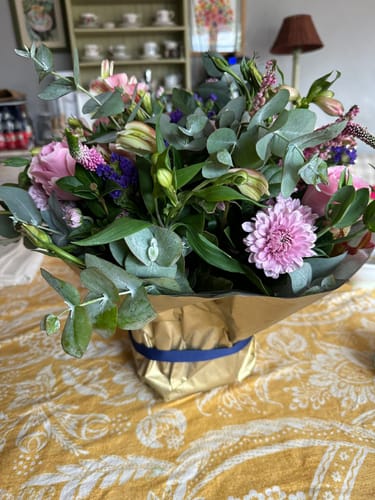 Customer's Harmony Flower Arrangement with fresh pink and purple flowers in a gold-wrapped container on a table.