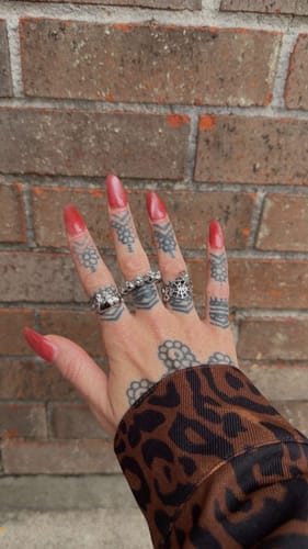 Reviewer's tattooed hand with red nails wearing the 10 Stages of Addiction Ring with other silver rings.