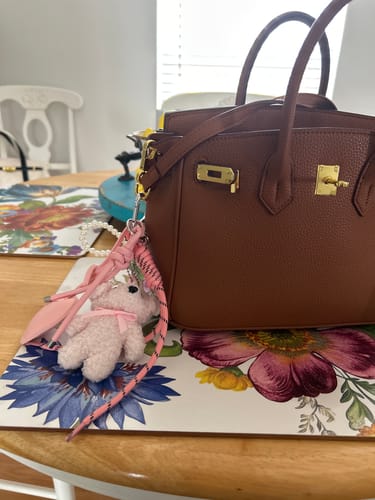 Customer's pink Bear Plush Bag Charm attached to the strap of a brown leather handbag, resting on a table.
