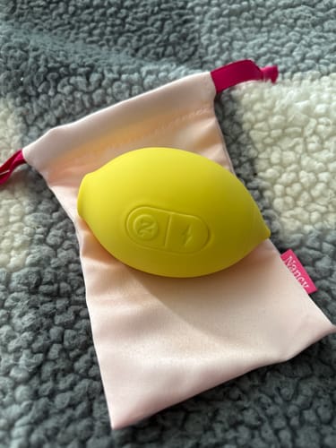 Reviewer's yellow Lem Clitoral Massager resting on its pink satin pouch on a fluffy gray blanket.
