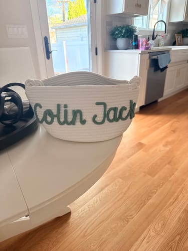 Customer's white personalized knitted basket with the name 'Colin Jack' in green yarn on a table.
