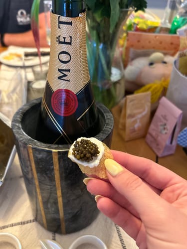Customer's hand holding a cracker with fresh caviar and cream, with a chilled bottle of champagne in the background.