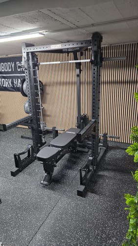 Ein Kunde zeigt sein hohes R7 Squat Rack in Schwarz, das in einem Fitnessraum mit einer Hantelbank aufgebaut ist.