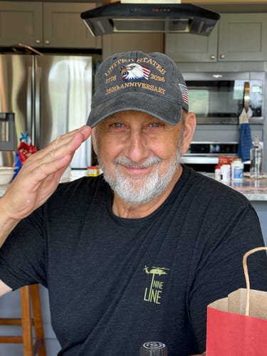 Reviewer wearing the embroidered USA 250th Anniversary Eagle Cap while smiling and saluting indoors.