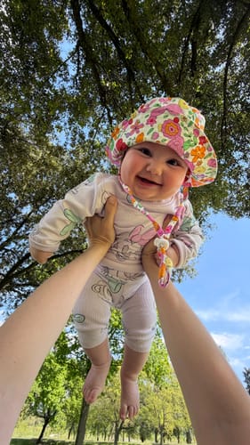 Customer's happy baby being held up outdoors, wearing the floral Paloma baby cotton hat.