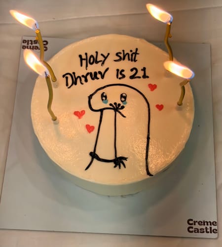 Reviewer's Meme Age Theme Cake, a round white cake with a crying meme, the text 'Holy shit Dhruv is 21', and four lit candles.