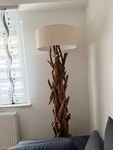 Rezensent zeigt die Stehlampe BLUMA aus Teakholz mit dem hellen, beigen Lampenschirm in einem Wohnzimmer.