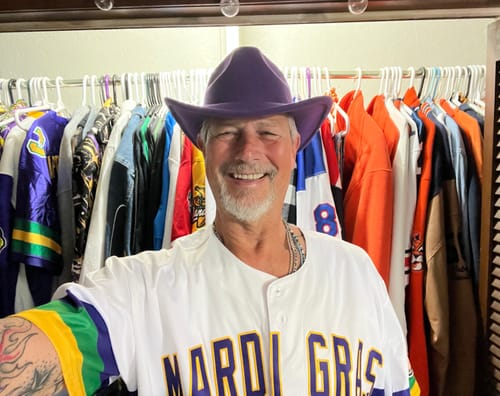 Customer smiling in a white Mardi Gras baseball jersey and purple hat, standing in front of a closet full of jerseys.