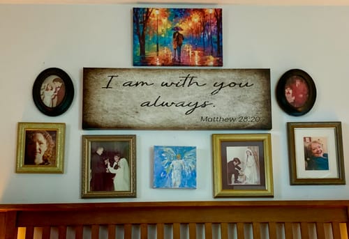Reviewer's 'I Am With You Always' sign displayed on a wall with framed photos above a bed.