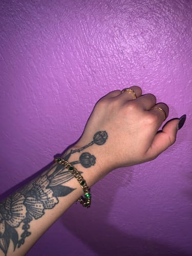 Cliente mostrando la Pulsera Kaffy dorada con piedras verdes en su muñeca contra una pared morada.