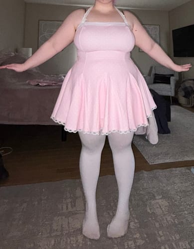Reviewer standing in a bedroom wearing the pink Dolly Polka Dot Dress with white halter straps and a full, flared skirt.
