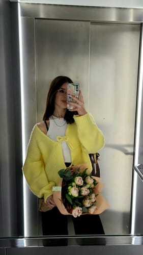 Kundin trägt den weichen, gelben Schleifen-Cardigan zu einem weißen Top und schwarzer Hose.