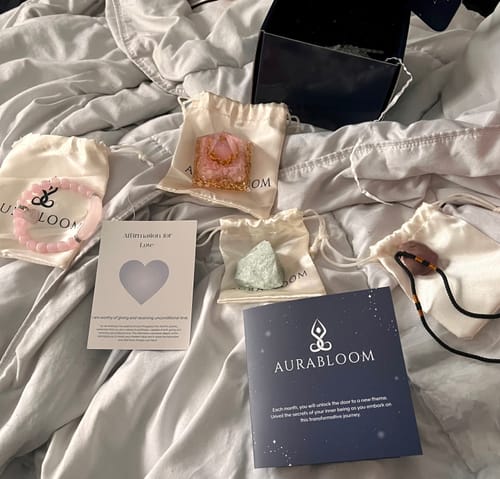 Customer shows the contents of their AuraBloom Subscription Box, including crystals and jewelry, laid out on a bed.