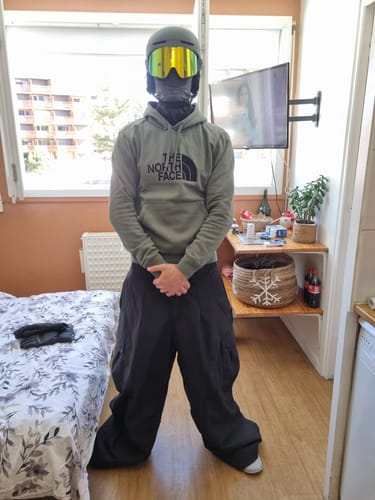 Reviewer showing the oversized fit of the black baggy snow pants while wearing them indoors with a hoodie and helmet.