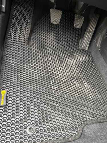 Recenzent pokazuje dopasowany czarny dywanik samochodowy zainstalowany w podłodze po stronie kierowcy.