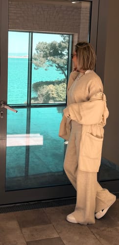 Kundin in einer beigen Essential Lounge Strickhose und passender Jacke blickt seitlich aus einem Fenster auf das Meer.