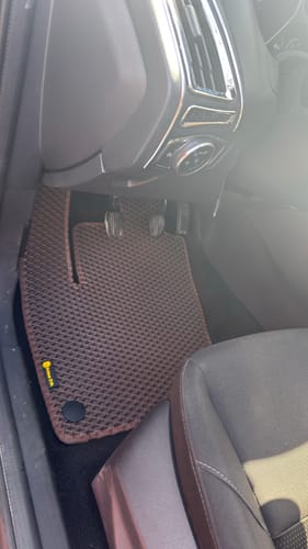 Recenzent pokazuje brązowy, dopasowany dywanik samochodowy po stronie kierowcy w Fordzie Focus.