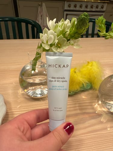 Kundens hand håller upp en tub Tiny Miracle Lips & Dry Spots framför ett bord.