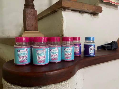 Cliente exibe vários potes do suplemento Happy Hair e os brindes Happy Sleep e Happy Collagen sobre uma superfície de madeira.