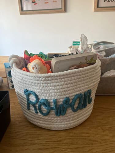 Customer's white personalized knitted basket with the name 'Rowan' in teal yarn, filled with baby items on a table.