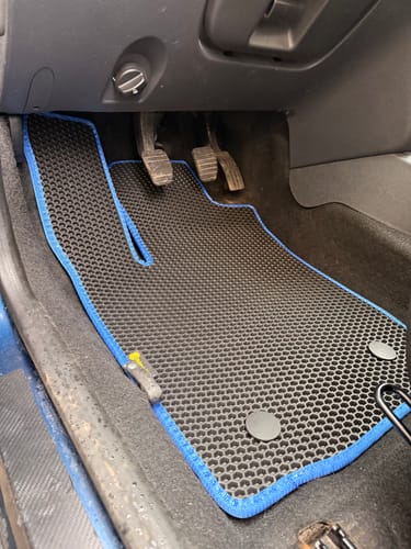 Recenzent pokazuje czarny dywanik samochodowy z niebieską obwódką, idealnie dopasowany do podłogi po stronie kierowcy.