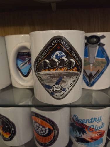 Customer's white Artemis II mug displayed on a glass shelf, showing the detailed mission patch design among other mugs.