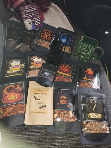 Kunde zeigt den Inhalt der Brew Mystery Box: zahlreiche Teesorten, eine grüne Tasse in Buchform und eine Duftlampe in Kesselform.