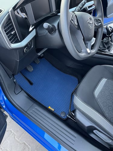 Recenzent pokazuje niebieski, dopasowany dywanik samochodowy po stronie kierowcy w nowoczesnym aucie.