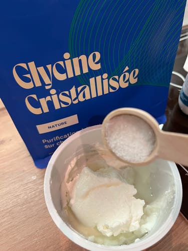 Un client ajoute une cuillère de Glycine Cristallisée dans un yaourt, avec le sachet du produit à l'arrière-plan.