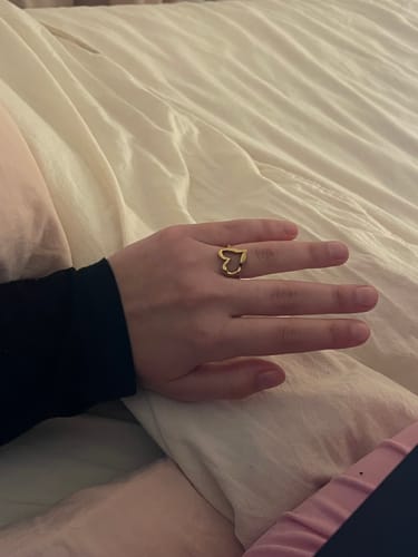 Customer's hand wearing the gold Eternal Heart Ring on their finger against white bedding.