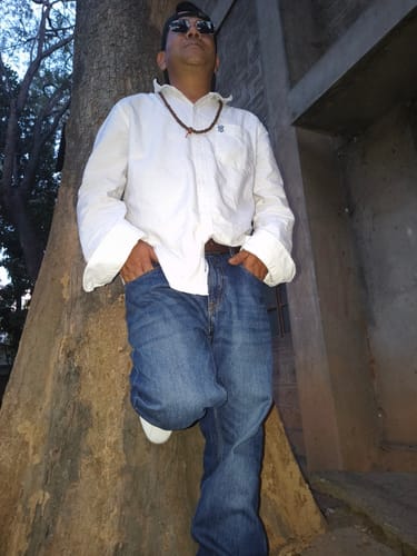 Reviewer models the Malana round sunglasses with a golden frame while leaning against a tree outdoors.