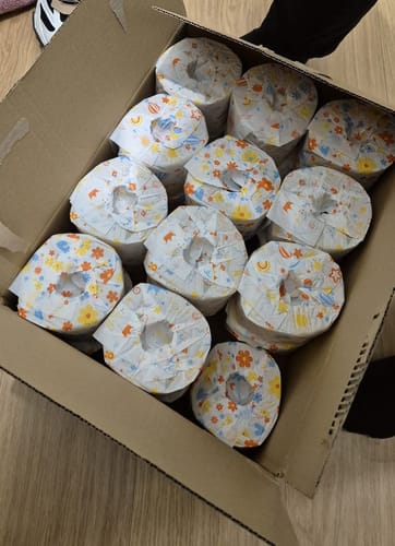 Un client montre une boîte en carton ouverte remplie de rouleaux de PAPIER TOILETTE Beau et Doux colorés.