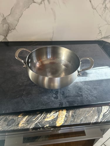 Rezensent zeigt den gehämmerten tiefen Titan-Topf auf einem dunklen Kochfeld in einer Küche.