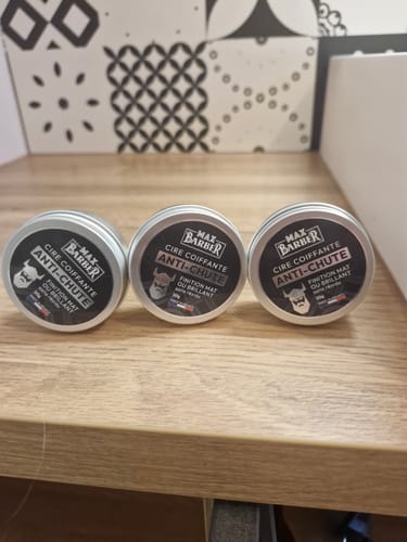 Un client montre trois pots de Cire Coiffante Anti-Chute alignés sur une étagère en bois.