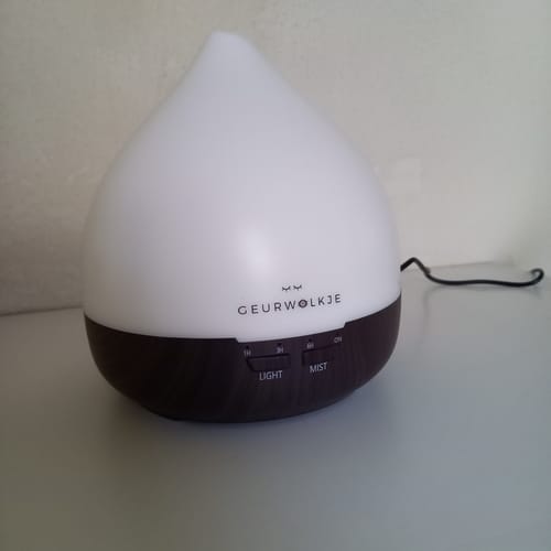 Recensent toont de Unity 2.0 aroma diffuser met een donkerhouten basis en een verlicht wit bovenstuk op een witte tafel.