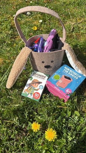 Ein Kunde zeigt das Spiel 'Gefunden! Outdoor Suchspiel' neben einem Korb in Hasenform und einem Kindermikroskop auf einer Wiese.