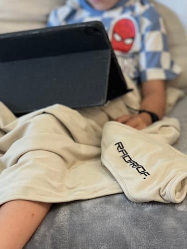 Customer's child using the Baby Blanket & Baby Beanie Bundle while relaxing on a couch with a tablet.