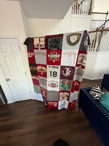 Customer holding up their finished Project Repat T-Shirt Quilt, made from red, grey, and white t-shirt squares in a home.